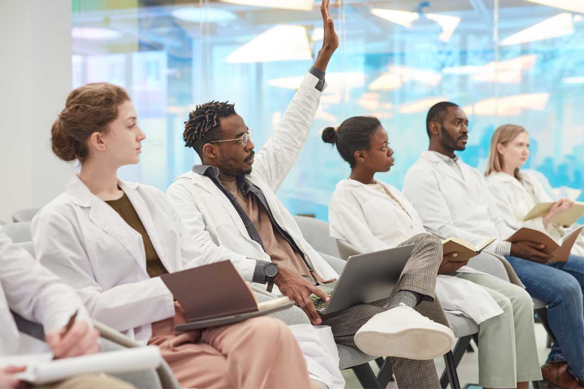 Group of individuals in lab coats seated in a classroom setting, with one person raising a hand to ask a question.
