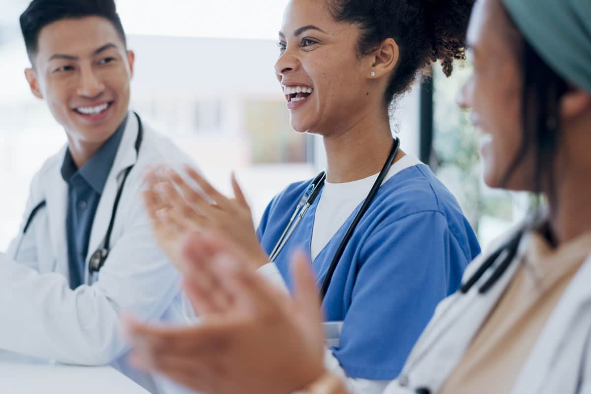 Three healthcare professionals in medical attire engaged in a discussion, smiling and clapping in a collaborative setting.