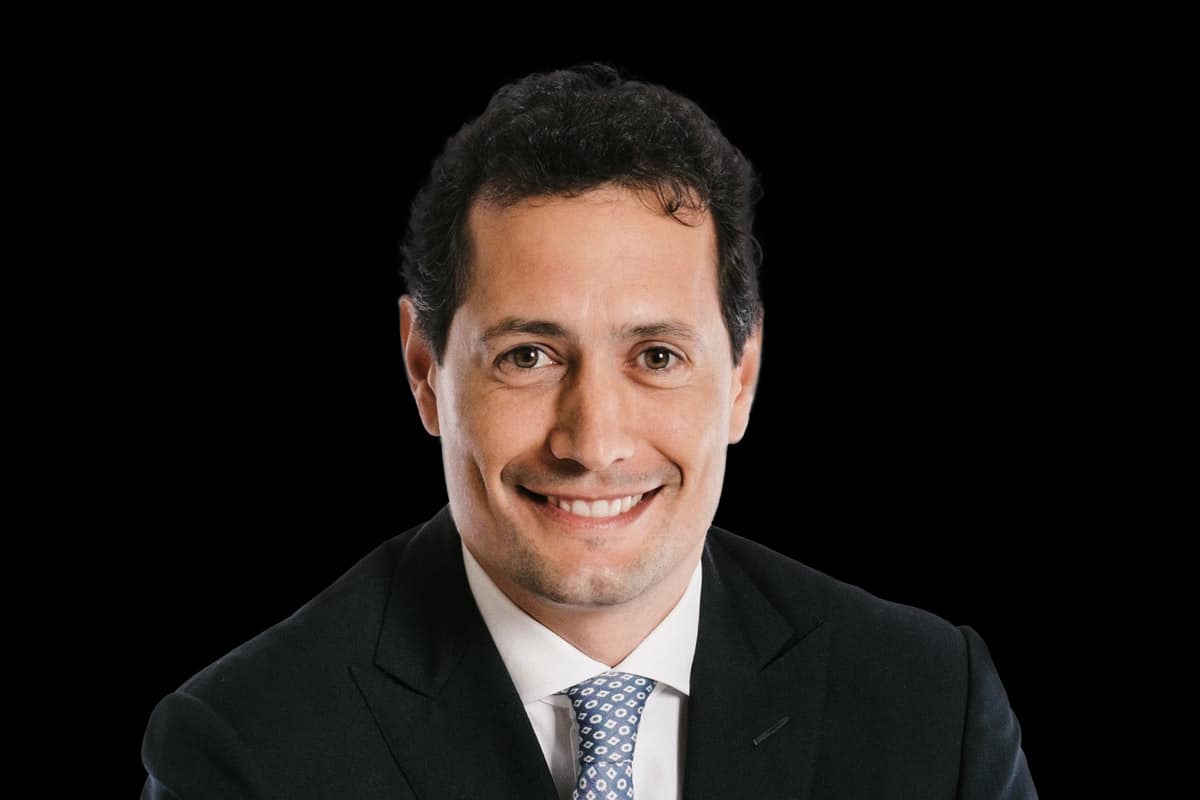 Portrait of a smiling man in a dark suit and light dotted tie against a solid black background; Tabanella conveys professionalism and confident presence.