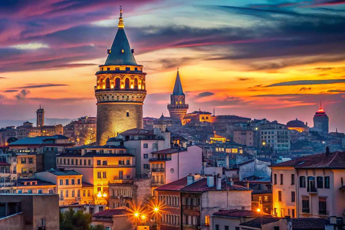 Sunset cityscape of Istanbul with the Galata Tower lit up over a dense historic district, warm orange and purple skies, and glowing streetlamps softly.