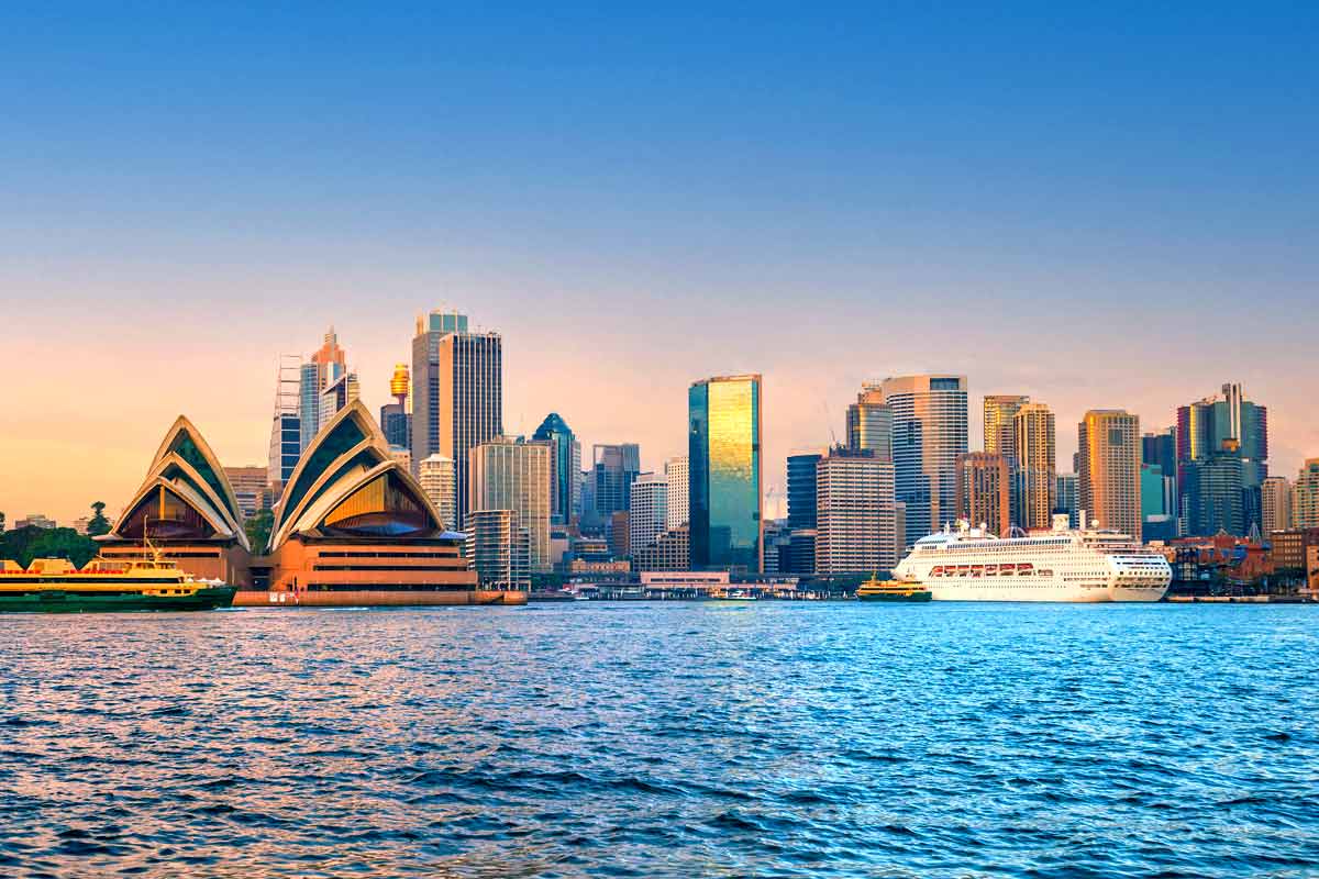 Sydney skyline at sunset featuring the Opera House and modern high-rises along the harbor, with blue water in the foreground and warm reflections.