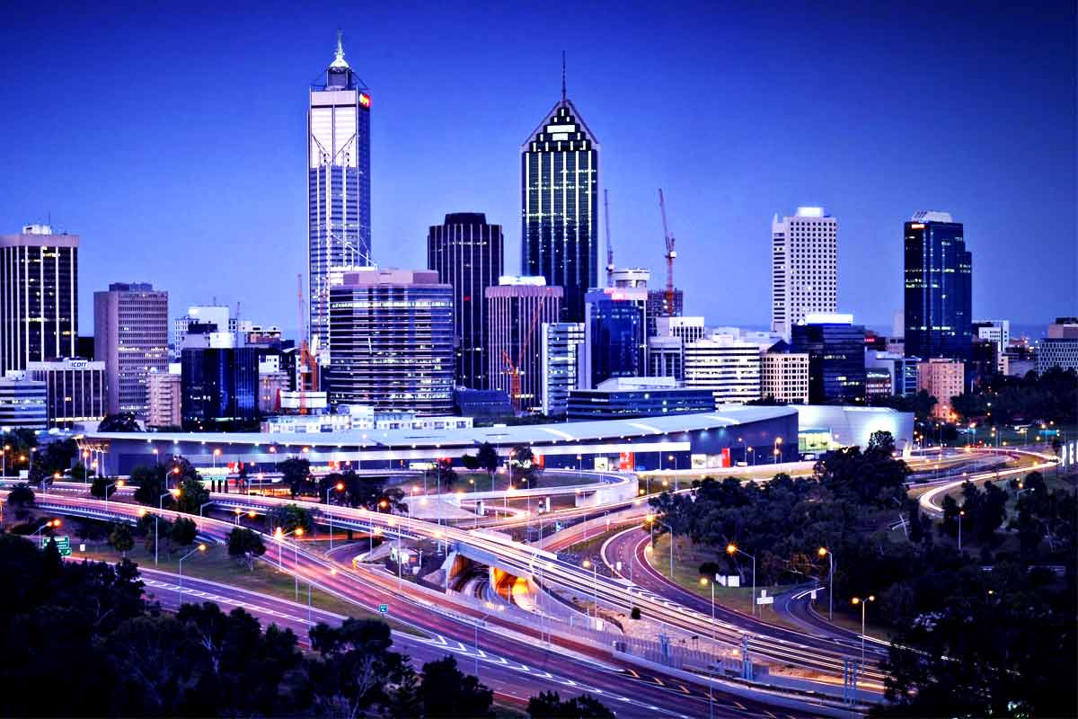 Perth city skyline at twilight with tall glass skyscrapers and winding highways, bathed in vibrant blue hues across the urban landscape.