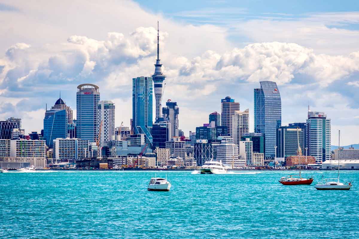 Auckland skyline featuring tall, glassy skyscrapers rises over a turquoise harbor as small boats glide by, framed by a bright, partly cloudy sky and distant hills