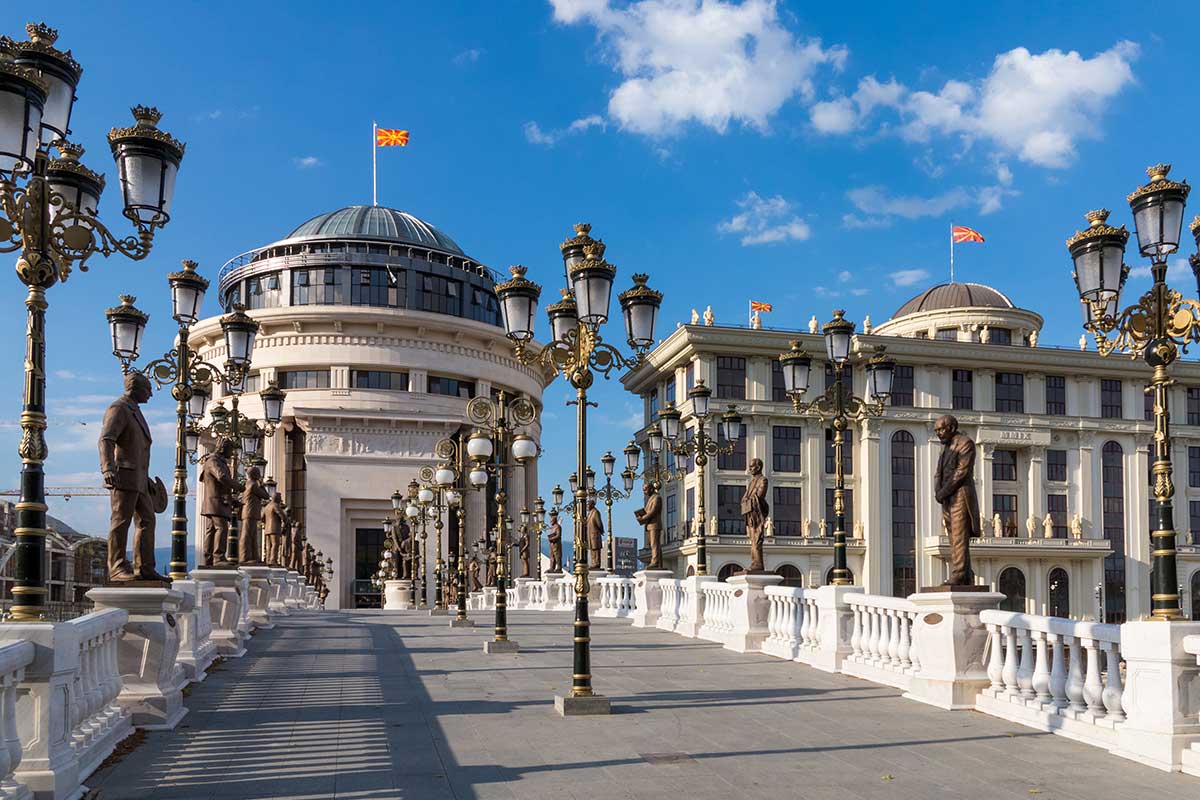 Skopje campus with domed neoclassical buildings, ornate street lamps, and bronze statues along a wide pedestrian promenade under a clear blue sky