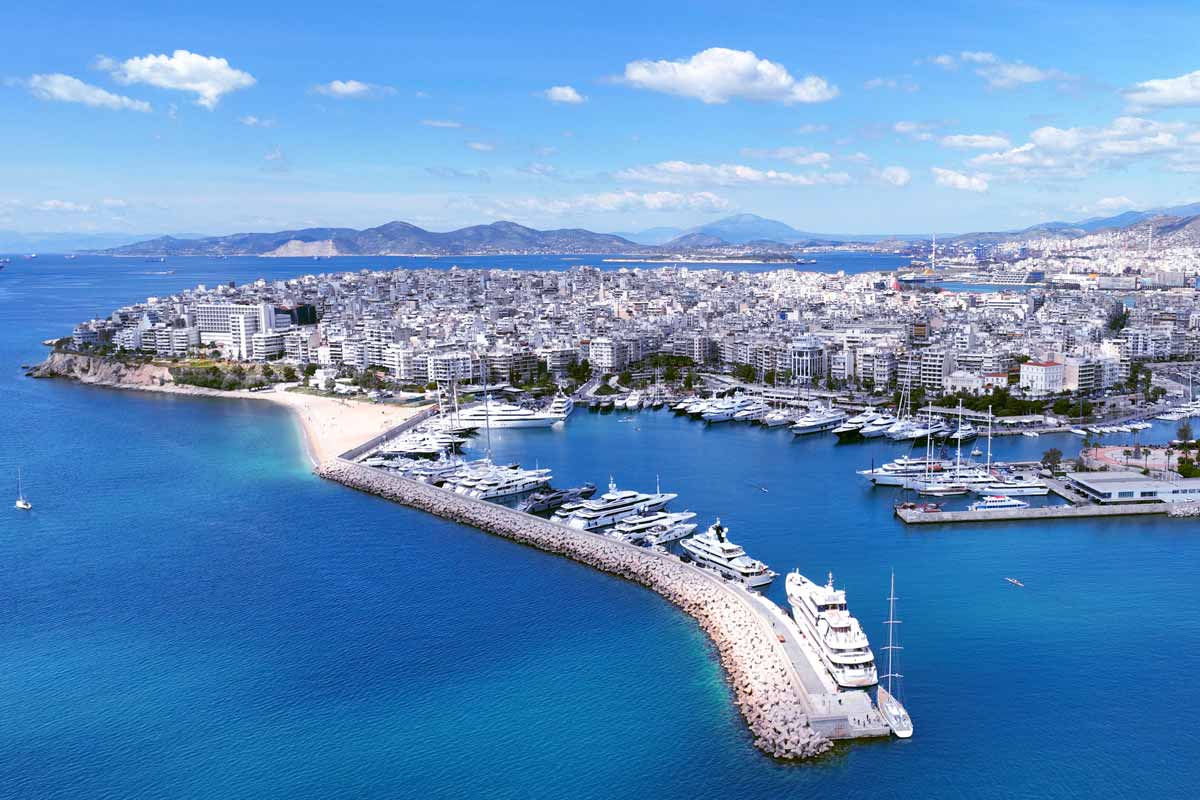 Aerial view of a sunny coastal harbor with a curved breakwater, blue water, and a marina filled with yachts beside a dense cityscape.