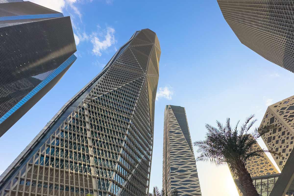 Riyadh skyline with tall glass skyscrapers rising against a blue sky and a palm tree in the foreground, emphasizing modern urban architecture and dramatic vertical lines.