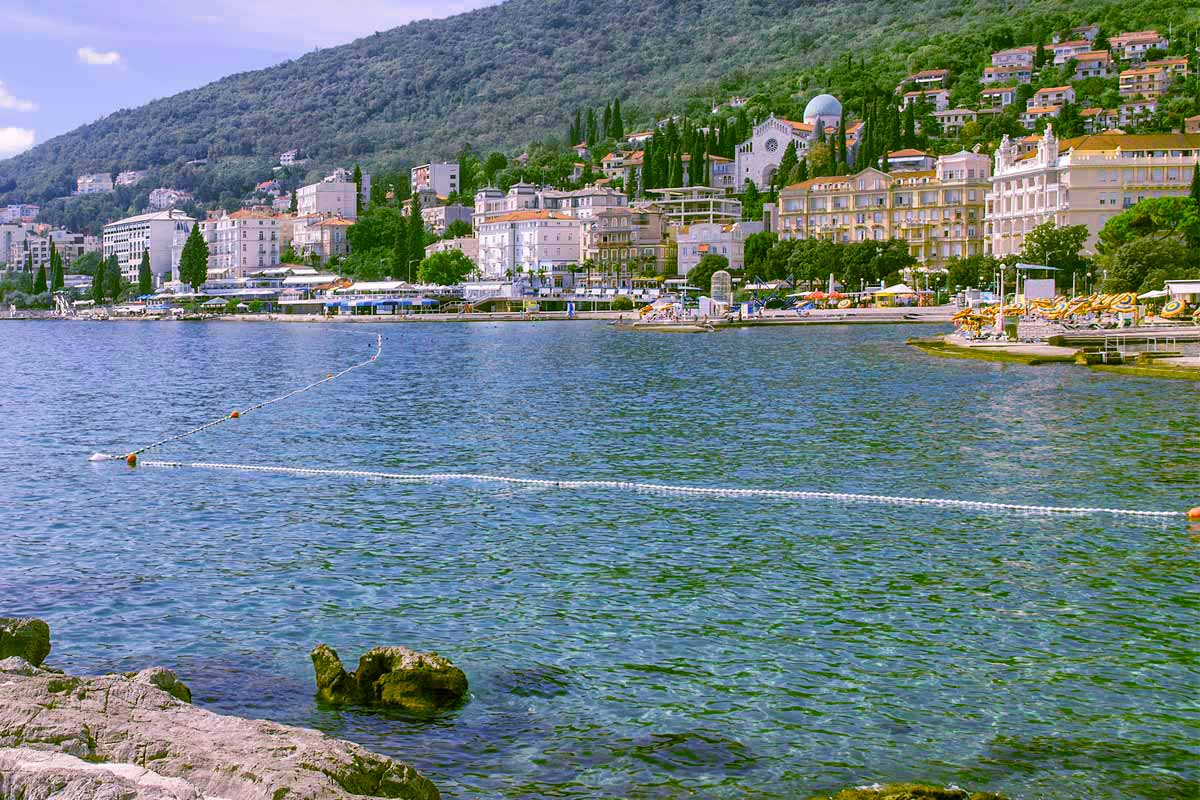 Opatija waterfront along the Adriatic Sea with blue water, rocky shore, and a hillside town of pastel buildings, trees, and seaside promenade.
