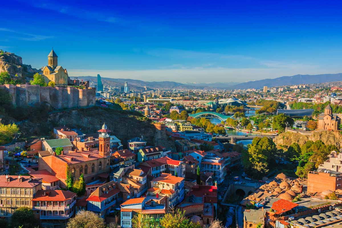 Tiblisi cityscape featuring ancient fortress walls on a hill, vibrant red-tiled rooftops, a winding river, and a modern skyline under a bright blue sky in Georgia.