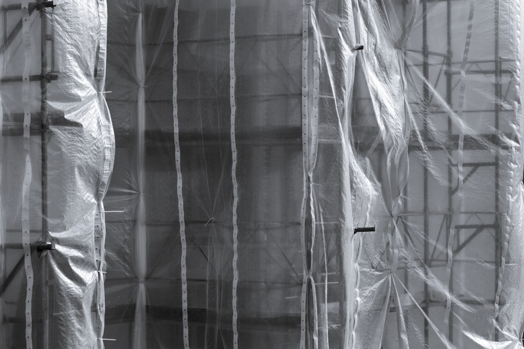 Gray construction scaffolding wrapped in translucent plastic sheeting, with metal poles and framework visible through the protective wrap, creating an industrial, textured scene.